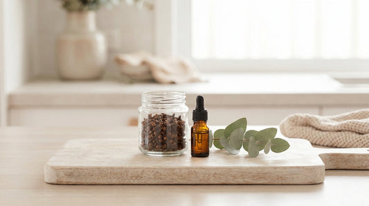 Whole cloves and a small amber dropper bottle on a pale timber board with a sprig of greenery, soft natural light.